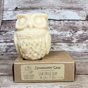 May include: A brown cardboard box with a white label that says "Cedarwood Sage Goat Milk Soap with Shea & Olive 3.5-4 oz" and a logo that says "Solace Farm Homestead". The box is sitting on a wooden surface and a white owl-shaped soap is sitting on top of the box.