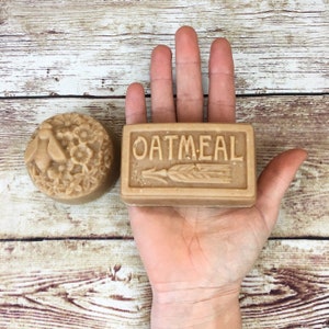 K&ouml;nnte beinhalten: Zwei handgemachte Seifen, eine mit einem Blumenmuster und die andere mit dem Wort "OATMEAL" und einem Weizen&auml;hren-Design. Die Seifen liegen auf einer Holzoberfl&auml;che.