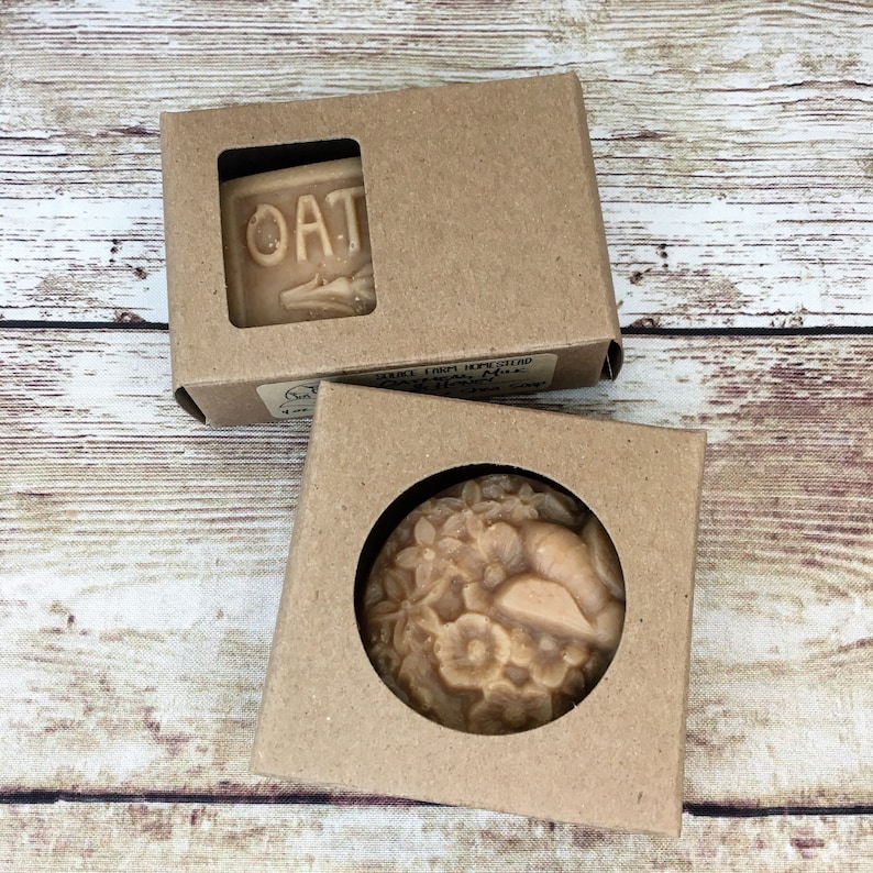 K&ouml;nnte beinhalten: Zwei braune Pappkartons mit ausgeschnittenen Fenstern, die handgemachte Seifenst&uuml;cke zeigen. Eine Schachtel zeigt eine Seife mit dem eingepr&auml;gten Wort "OAT". Die andere Schachtel zeigt eine Seife mit einem Blumenmuster und einer Schnecke.