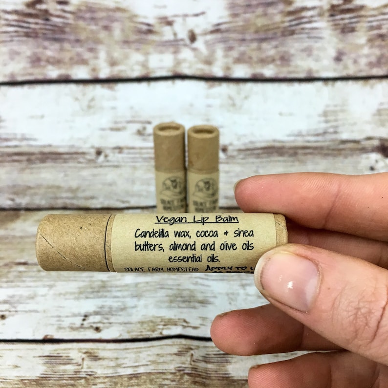 May include: A brown cardboard tube with a white label that reads "Vegan Lip Balm Candelilla wax, cocoa + shea butters, almond and olive oils essential oils. SOLACE FARM HOMESTEAD APPLY TO L"