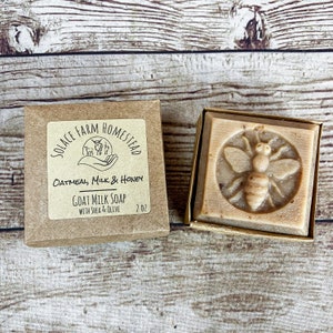 K&ouml;nnte beinhalten: Ein brauner Karton mit einem wei&szlig;en Etikett, auf dem "SOLACE FARM HOMESTEAD" und "OATMEAL, MILK & HONEY GOAT MILK SOAP WITH SHEA & OLIVE 2 OZ" steht. Die Schachtel enth&auml;lt eine Seife mit einem Bienenmotiv.
