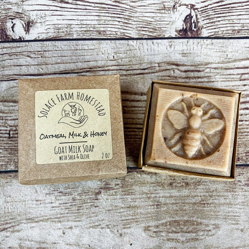 K&ouml;nnte beinhalten: Ein brauner Karton mit einem wei&szlig;en Etikett, auf dem "SOLACE FARM HOMESTEAD" und "OATMEAL, MILK & HONEY GOAT MILK SOAP WITH SHEA & OLIVE 2 OZ" steht. Die Schachtel enth&auml;lt eine Seife mit einem Bienenmotiv.