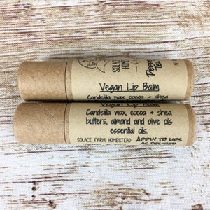 May include: Two brown cardboard tubes with labels for vegan lip balm. The labels list the ingredients: candelilla wax, cocoa, shea butters, almond and olive oils, essential oils. The bottom label also says "Apply to lips as desired" and "Solace Farm Homestead".