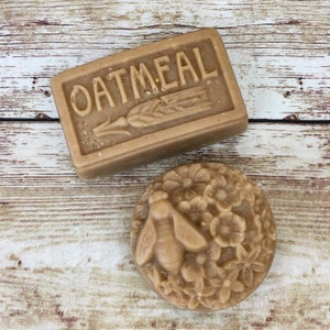 K&ouml;nnte beinhalten: Zwei handgemachte Seifenst&uuml;cke. Ein St&uuml;ck ist rechteckig und hat das Wort "OATMEAL" gepr&auml;gt. Das andere St&uuml;ck ist rund und hat eine Biene und Blumen gepr&auml;gt.