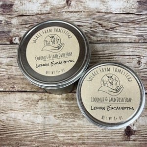 May include: Two metal tins of coconut and lard dish soap with a brown paper label. The label says "Solace Farm Homestead" and "Coconut & Lard Dish Soap Lemon Eucalyptus". The smaller tin is labeled "Net Wt 8 oz" and the larger tin is labeled "Net Wt 14 oz".