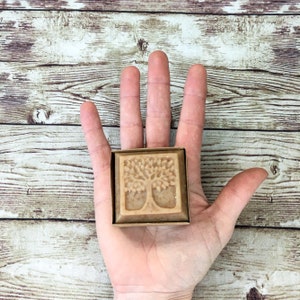 K&ouml;nnte beinhalten: Eine quadratische Seife mit einem Baumdesign auf der Vorderseite. Die Seife befindet sich in einer Pappbox und wird in der Hand einer Person gehalten.