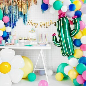 May include: A birthday party table setting with a green cactus balloon, a white daisy balloon, and a cake with a floral design. The table is decorated with colorful balloons, plates, and cups. The banner says "Happy Birthday".