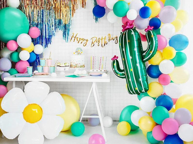 May include: A birthday party table setting with a green cactus balloon, a white daisy balloon, and a cake with a floral design. The table is decorated with colorful balloons, plates, and cups. The banner says "Happy Birthday".