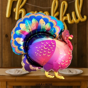 May include: A colourful turkey-shaped balloon with a vibrant peacock-inspired tail. The balloon features shades of blue, purple, pink, and gold. The turkey has gold feet and a red wattle. The balloon is likely for Thanksgiving.