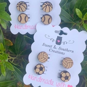 May include: A set of six wooden earrings featuring different sports ball designs. The earrings are displayed on two white cards with the text "Sweet & Southern Creations" and "Handmade with" on them. The earrings are made of wood and have a natural finish.