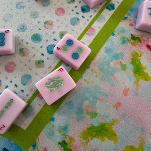 May include: Several light pink Mahjong tiles featuring engraved symbols and numbers in blue, green, and red ink. The tiles display various markings including the letter F, the number 4, and circular patterns, scattered across a colorful patterned surface.
