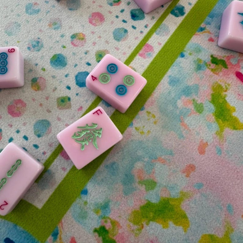 May include: Several light pink Mahjong tiles featuring engraved symbols and numbers in blue, green, and red ink. The tiles display various markings including the letter F, the number 4, and circular patterns, scattered across a colorful patterned surface.