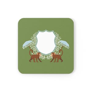 Puede incluir: Posavasos cuadrado con fondo verde, con un diseño blanco en forma de escudo con detalles florales. Dos monos marrones con paraguas azules flanquean el escudo. Un artículo decorativo.