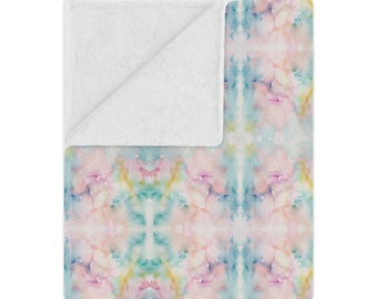 Op de afbeelding: Een rechthoekige plaid met een zacht, op aquarel geïnspireerd patroon met gemengde tinten pastelroze, lichtblauw en lichtgeel. De bovenhoek is omgeslagen om een effen witte, getextureerde, fleece-achtige onderkant te onthullen.