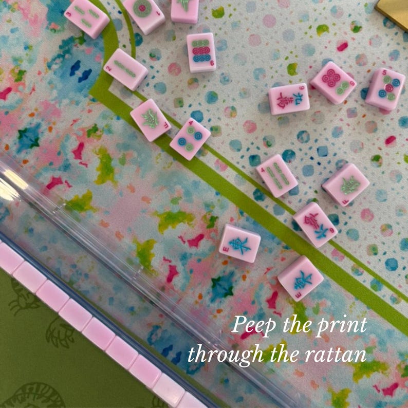 May include: A collection of small, rectangular, light pink Mahjong tiles featuring various colorful symbols and numbers, scattered across a surface with a watercolor-style pattern of blue, green, and pink, with the text Peep the print through the rattan.