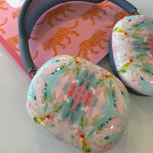 May include: Over-ear headphones featuring a grey headband and ear cups with a glossy, multi-colored abstract paint finish in shades of pink, light blue, and yellow. The headphones are positioned over a pink surface with an orange tiger pattern.