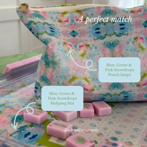 May include: A fabric pouch with a blue, green, and pink floral pattern and a gold zipper, positioned above a matching mat featuring the same pattern. Several small, rectangular, light pink mahjong tiles are scattered across the mat surface.