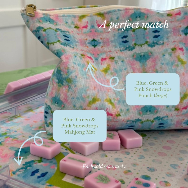 May include: A fabric pouch with a blue, green, and pink floral pattern and a gold zipper, positioned above a matching mat featuring the same pattern. Several small, rectangular, light pink mahjong tiles are scattered across the mat surface.