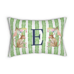 May include: Rectangular decorative pillow with a green and white striped background. The pillow features two watercolor monkeys holding umbrellas, with the letter "E" in navy blue in the center. The monkeys are light brown.