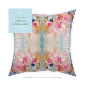 May include: A square throw pillow featuring an abstract pattern with blended strokes of pink, orange, cream, and light blue. The design is symmetrical, mirroring itself from the center. A light blue square overlay contains the text Elizabeth Mae & Co.