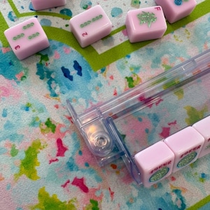 May include: A set of light pink Mahjong tiles with green and red markings, including tiles labeled Joker, arranged on a clear plastic rack. The tiles feature various symbols and numbers, with some scattered on a colorful, watercolor-patterned surface.
