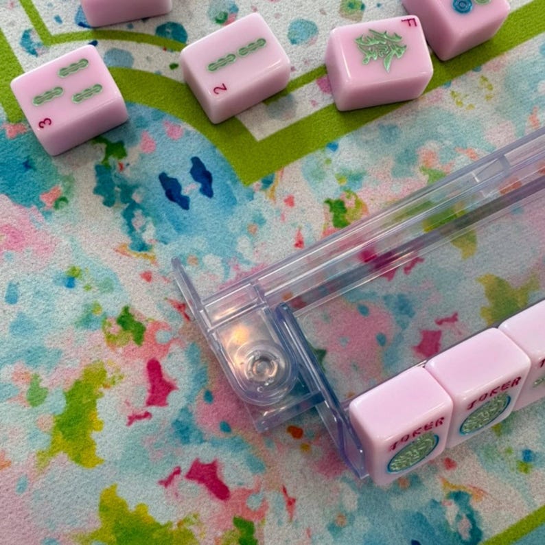 May include: A set of light pink Mahjong tiles with green and red markings, including tiles labeled Joker, arranged on a clear plastic rack. The tiles feature various symbols and numbers, with some scattered on a colorful, watercolor-patterned surface.