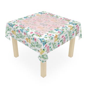 May include: A square white tablecloth with a colorful floral and pagoda design. The pattern includes pink, blue, yellow, and green elements. The tablecloth is draped over a small wooden table with four legs.