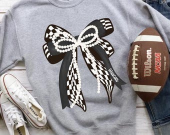 Peut inclure: Un sweat-shirt gris avec un grand nœud à carreaux noirs et blancs orné de perles. Un ballon de football américain marron et des baskets blanches sont également présents. Le mot "Wildcats" est sur le nœud.