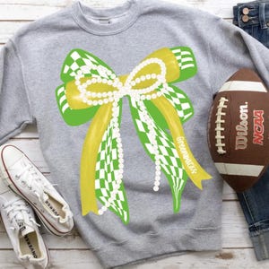 Op de afbeelding: Een grijze sweatshirt met een groot strikdesign in geel, groen en wit geblokte patronen, versierd met een parelketting. Een bruine voetbal en witte sneakers zijn ook zichtbaar.