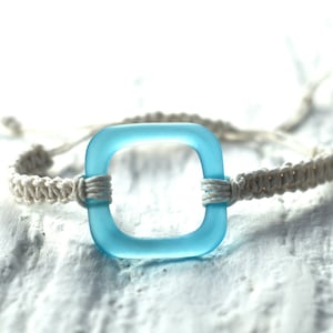 May include: A light blue square-shaped glass bead is woven into a white braided cord bracelet.