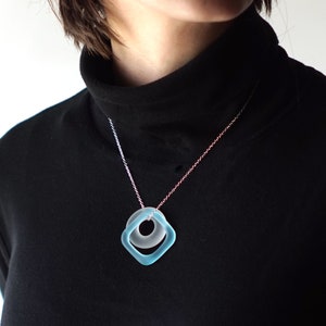 May include: A silver chain necklace with a light blue glass pendant featuring a square shape with a circle inside.