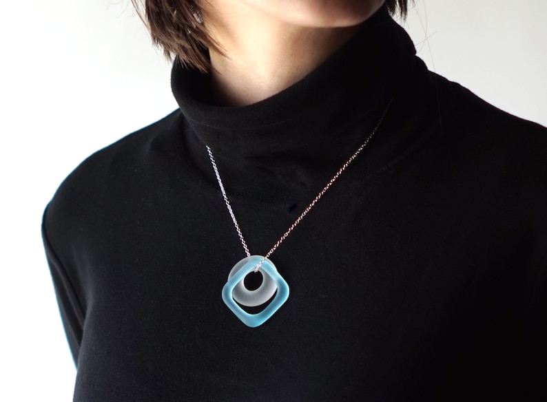 May include: A silver chain necklace with a light blue glass pendant featuring a square shape with a circle inside.