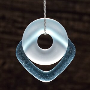 May include: A silver chain necklace with a light blue glass pendant. The pendant is a circle with a smaller circle cut out of the center, and it is surrounded by a larger, teardrop-shaped piece of glass.