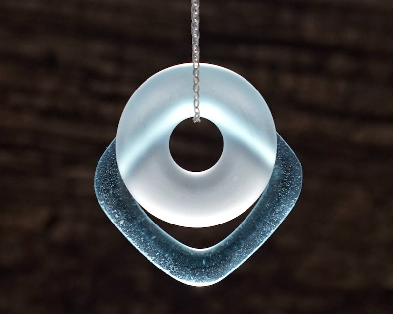 May include: A silver chain necklace with a light blue glass pendant. The pendant is a circle with a smaller circle cut out of the center, and it is surrounded by a larger, teardrop-shaped piece of glass.