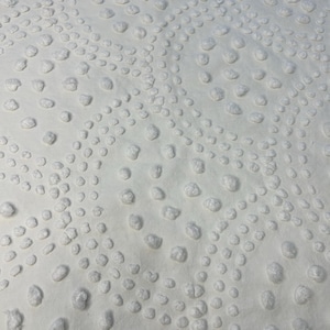May include: A white fabric featuring a raised, tufted texture. The surface is decorated with a repeating pattern of small, soft, rounded bumps arranged in curved, circular, and swirling lines that create a tactile, dimensional design.
