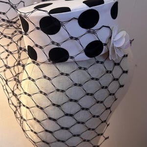 Könnte beinhalten: Ein weißer Hut mit großen schwarzen Polka Dots, verziert mit einem schwarzen Netzschleier und einer weißen Blume mit schillernden Akzenten. Der Hut wird auf einem Schaufensterkopf präsentiert.