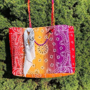 Könnte beinhalten: Eine farbenfrohe Tragetasche mit Patchwork-Design. Die Tasche zeigt ein rotes, orangefarbenes, gelbes und violettes Paisley-Muster. Ein weißes Bandana und eine Perlenkette hängen an den Griffen. Die Tasche ist aus gestepptem Stoff gefertigt.