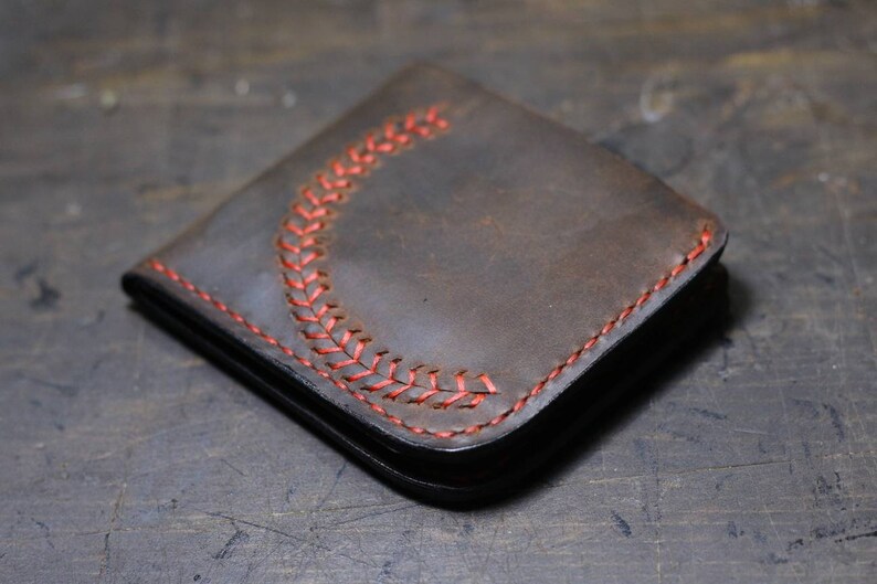 Op de afbeelding: Een bruine leren portemonnee met rode stiksels in de vorm van een honkbal-steekpatroon.