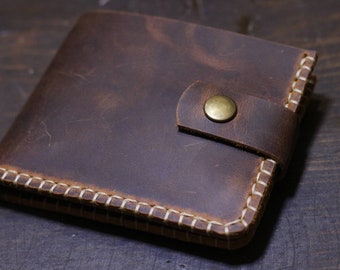 May include: A brown leather wallet with a flap closure and a brass button. The wallet has white stitching around the edges.