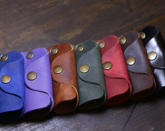 May include: A set of eight leather keychains in various colours, including blue, purple, brown, green, red, brown, black, and brown. Each keychain has a metal button closure.
