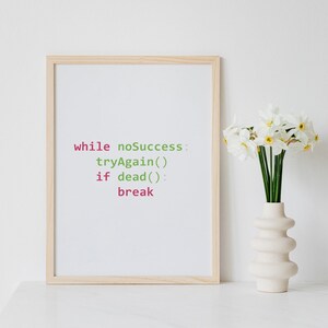 May include: A framed print with a light blue background and a code snippet in green and pink text that reads "while noSuccess: tryAgain() if dead(): break". The print is in a light wood frame and is sitting on a white surface next to a white vase with white daffodils.