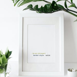 K&ouml;nnte beinhalten: Wei&szlig;er gerahmter Druck mit dem Text ".im-an-introvert { border-style: solid; }" in einer codeartigen Schriftart. Der Druck liegt auf einer wei&szlig;en Oberfl&auml;che mit gr&uuml;nen Pflanzen auf beiden Seiten.