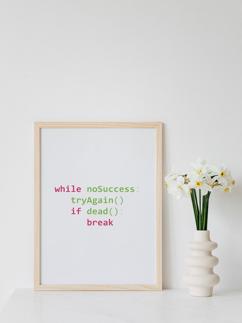 May include: A framed print with a light blue background and a code snippet in green and pink text that reads "while noSuccess: tryAgain() if dead(): break". The print is in a light wood frame and is sitting on a white surface next to a white vase with white daffodils.