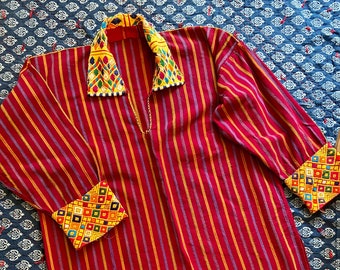 Puede incluir: Una camisa de manga larga a rayas rojas con cuello y puños decorativos. El cuello y los puños presentan un colorido estampado geométrico con cuadrados amarillos, verdes, azules y rojos. Una cadena dorada cuelga del cuello.