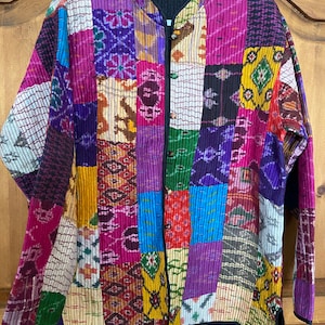May include: A colorful patchwork quilted jacket with a mandarin collar and button closure. The jacket is made of a variety of fabrics with different patterns and colors, including pink, purple, blue, green, yellow, and brown.