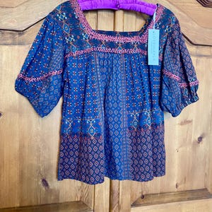 May include: A blue and red patterned blouse with a square neckline and short sleeves. The blouse has a crocheted trim around the neckline and sleeves. The tag on the blouse reads "Love + Kyla".