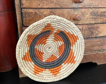 May include: A round woven basket with a natural straw base and orange, purple, and black geometric patterns.