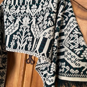 May include: A woven textile with a repeating pattern of white floral and geometric shapes on a dark green and cream background. The textile has a fringed edge and is hanging on a wooden door.