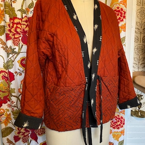 May include: An orange quilted jacket with a black patterned trim and tie closure. The jacket features a diamond pattern and a geometric design at the bottom. The sleeves and collar are trimmed with a black and white pattern. A unique, handcrafted garment.