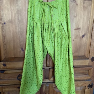 May include: Bright green pants with a drawstring waist and a loose, flowing fit. The fabric is adorned with small, evenly spaced gold-colored polka dots. The pants are hanging on a black hanger against a wooden background.